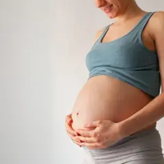 Uśmiechnięta ciężarna kobieta trzyma dłonie na brzuchu. Czas na poród zbliża się, może już niedługo pojawi się czop śluzowy.