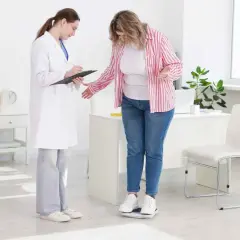 Lekarz obesitolog notuje wyniki ważenia pacjentki. Kobieta stoi na wadze, lekarz zapisuje dane.