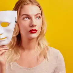 Blondynka z czerwoną szminką trzyma białą maskę, jakby zastanawiała się, czy być ekstrawertykiem, czy ukryć swoją prawdziwą twarz.