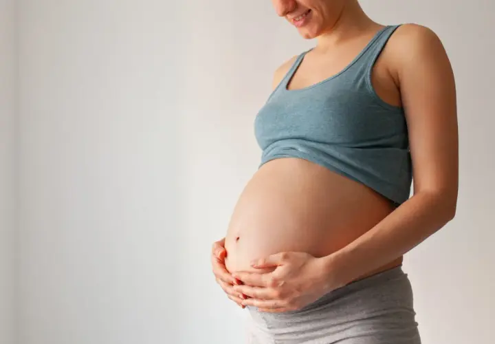 Uśmiechnięta ciężarna kobieta trzyma dłonie na brzuchu. Czas na poród zbliża się, może już niedługo pojawi się czop śluzowy.