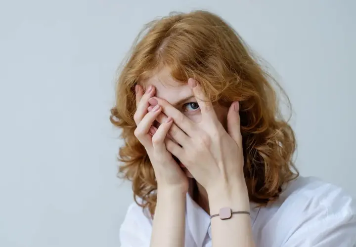 Rudowłosa dziewczyna z niebieskimi oczami zakrywa twarz dłońmi, jakby walczyła z fobią.