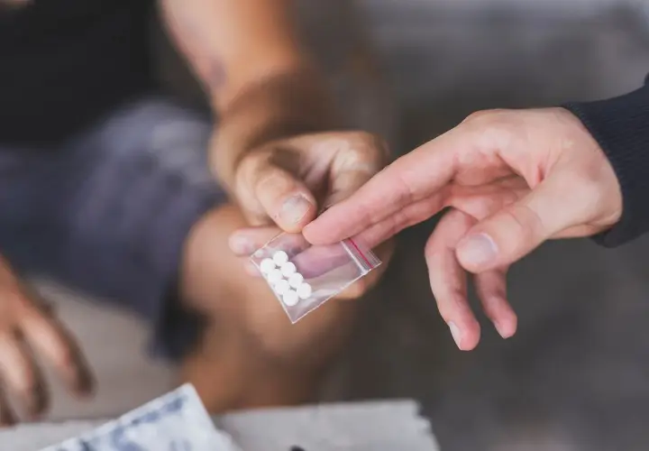 Dwie dłonie, jedna podaje, druga odbiera małą torebkę z białymi tabletkami, być może mdma.