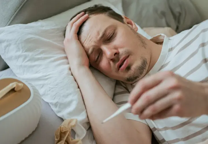 Chory mężczyzna leży w łóżku, trzymając termometr. Widać jego cierpiącą twarz i rękę na czole. To typowe objawy grypy wariant k.
