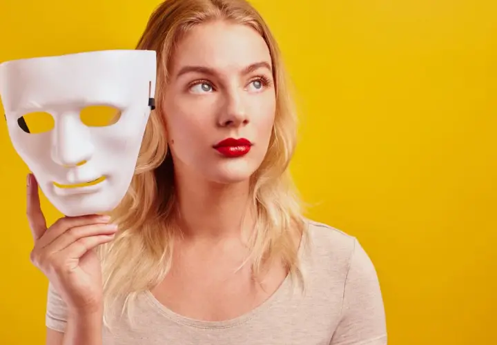 Blondynka z czerwoną szminką trzyma białą maskę, jakby zastanawiała się, czy być ekstrawertykiem, czy ukryć swoją prawdziwą twarz.