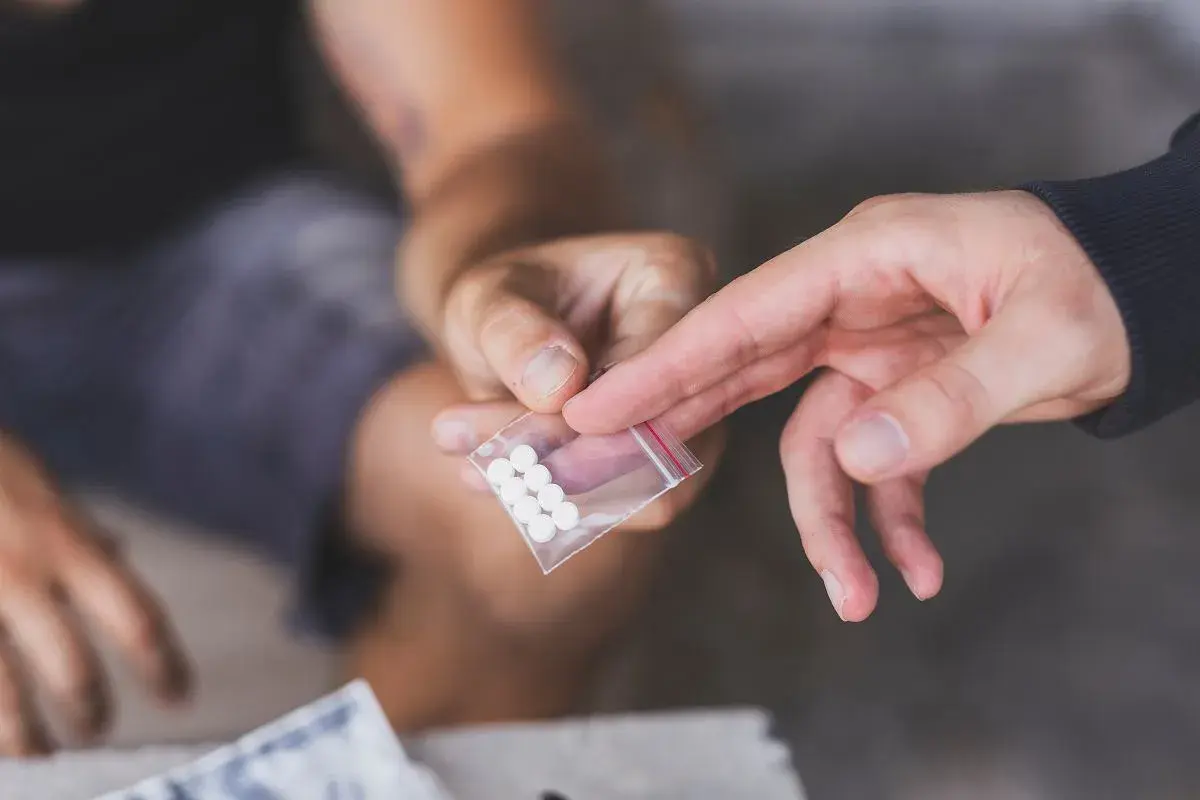 Dwie dłonie, jedna podaje, druga odbiera małą torebkę z białymi tabletkami, być może mdma.